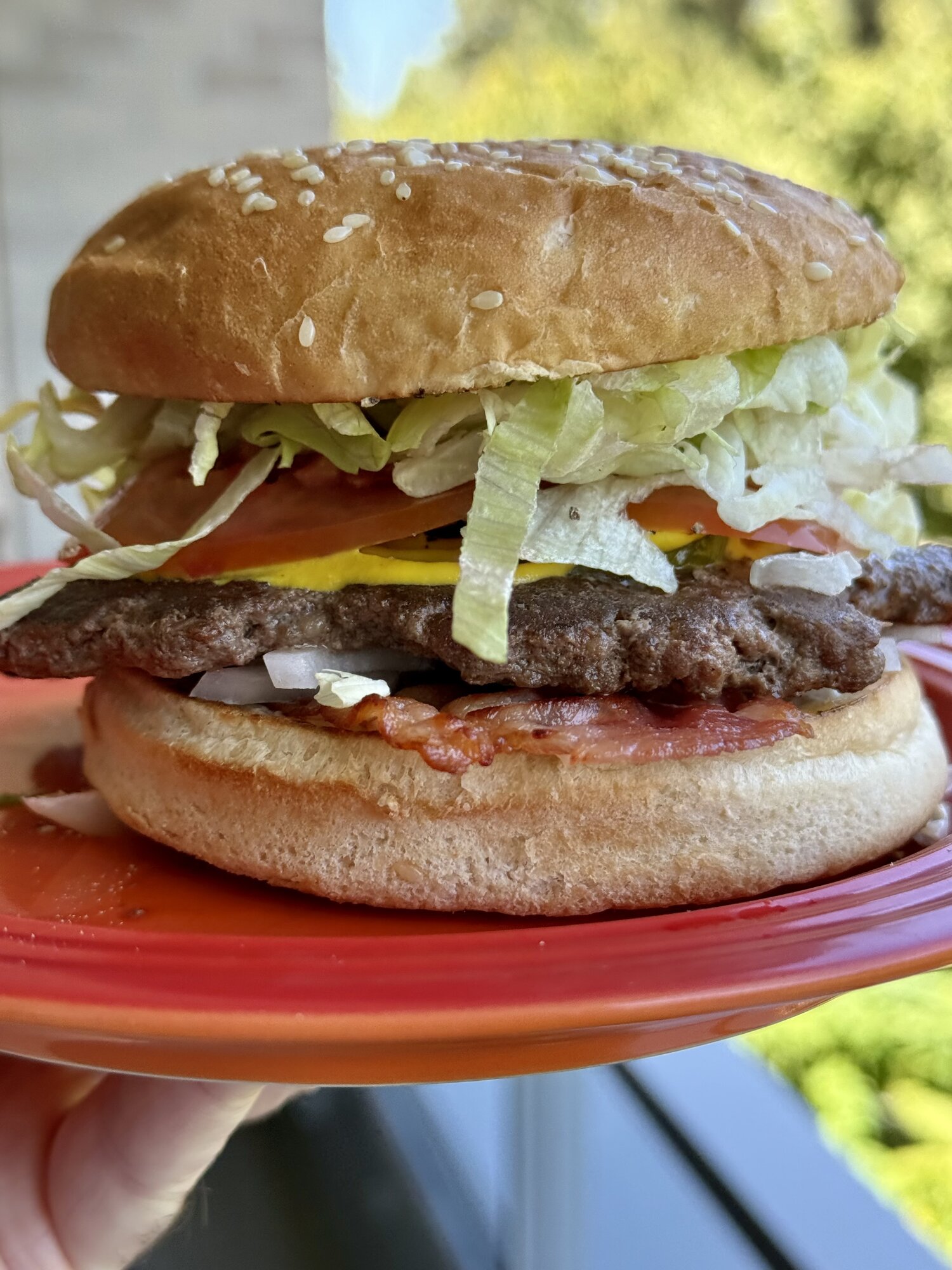 Burger with bacon, onion, mustard, tomato, and shredded lettuce, held up on a red plate