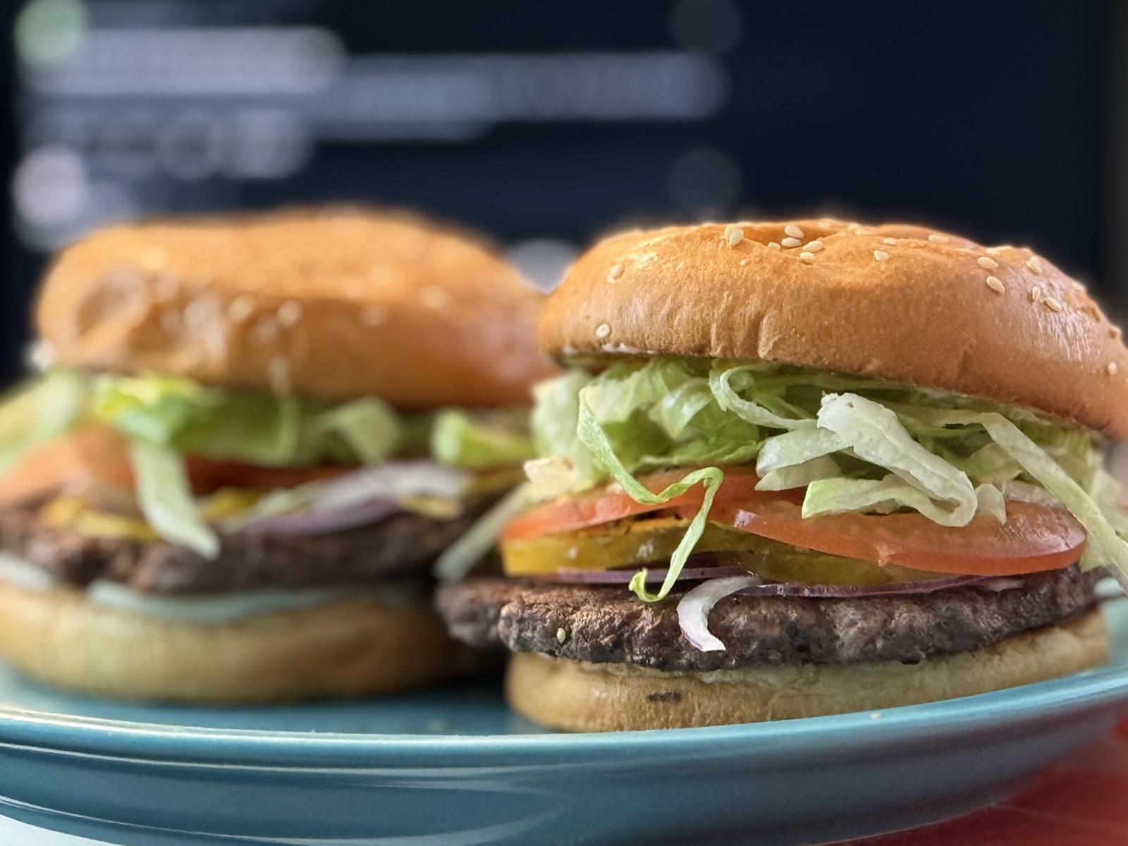 Two burgers with sesame buns, shredded lettuce, tomato, pickles, and red onion on a teal plate
