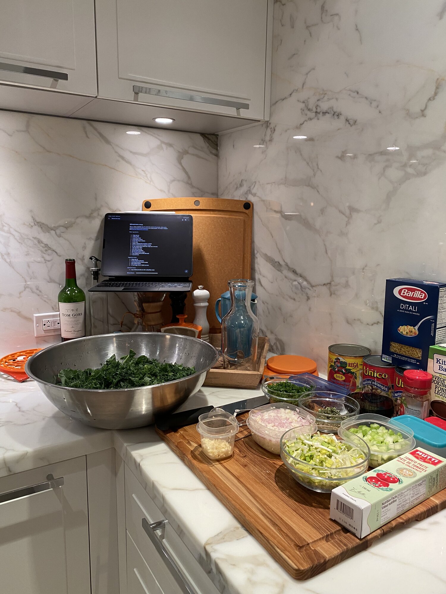 Mise en place for minestrone on a marble counter: kale, diced vegetables, canned tomatoes and beans, ditali, and the recipe on a tablet