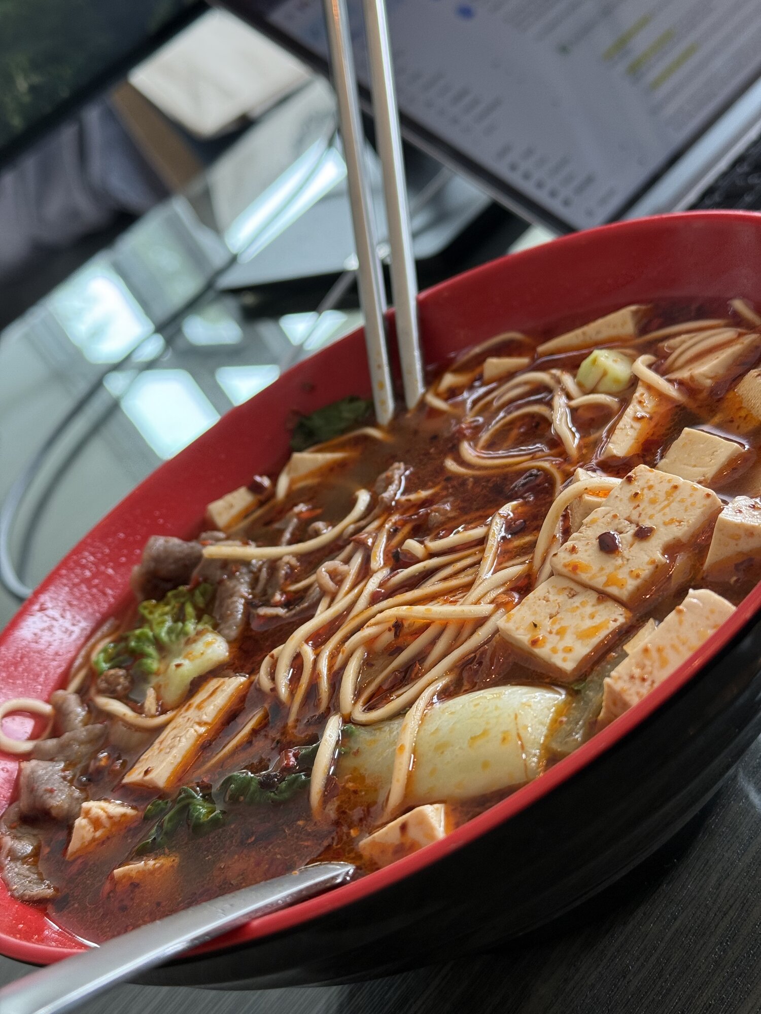 Spicy beef noodle soup with tofu, napa cabbage, and chili oil in a red bowl, chopsticks resting in it